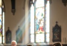 Cold stone pressed my cheek when I first learned what “night” meant. I couldn’t see the church doors—only hear them. A bell rang. Footsteps stopped. “Father… there’s a baby,” someone whispered. Then his voice, calm but shaken: “Who leaves a child in God’s cold?” I reached out into darkness and grabbed air. “Don’t go,” I cried—though I didn’t know who I was begging. Years later, the priest’s hands guided mine to a candle flame. “Light isn’t only seen,” he said. I laughed… until I heard a woman scream my name from the altar.